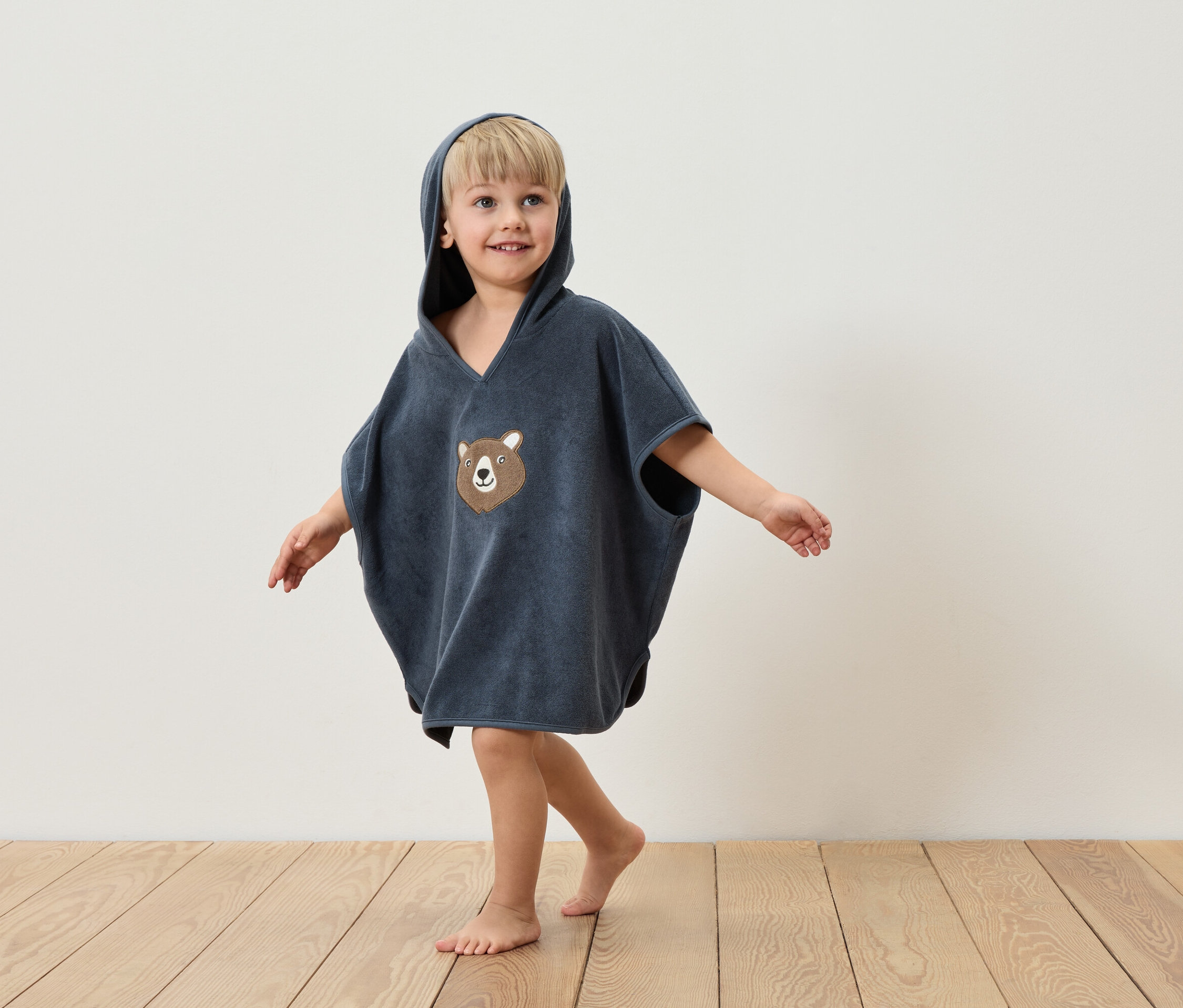 Ein Junge in einem blauen Frottee-Poncho mit Kapuze und Bärenapplikation steht auf einem Holzboden vor einer weißen Wand.