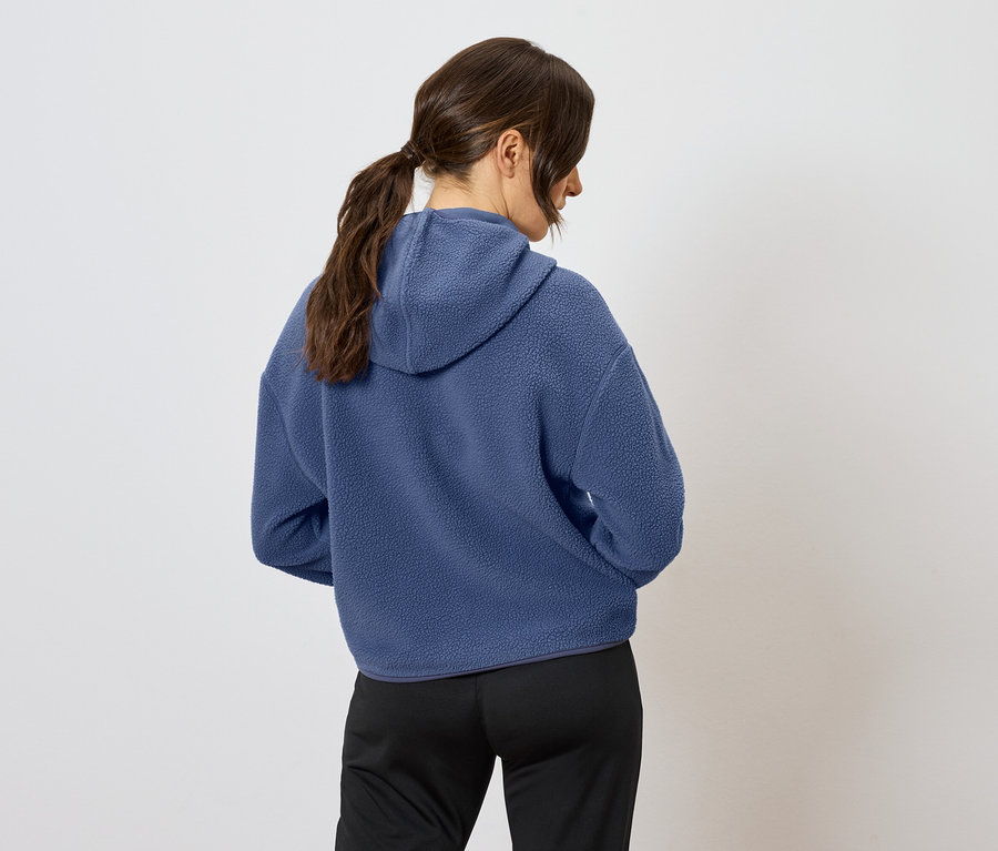 Rückansicht einer Frau mit braunen, zum Pferdeschwanz zusammengebundenen Haaren, die einen blauen Hoodie und eine schwarze Hose trägt.