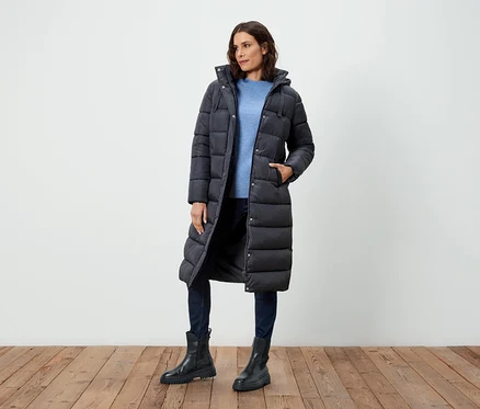 Frau posiert in einem langen, schwarzen Daunenmantel, blauem Pullover, Jeans und schwarzen Stiefeln.