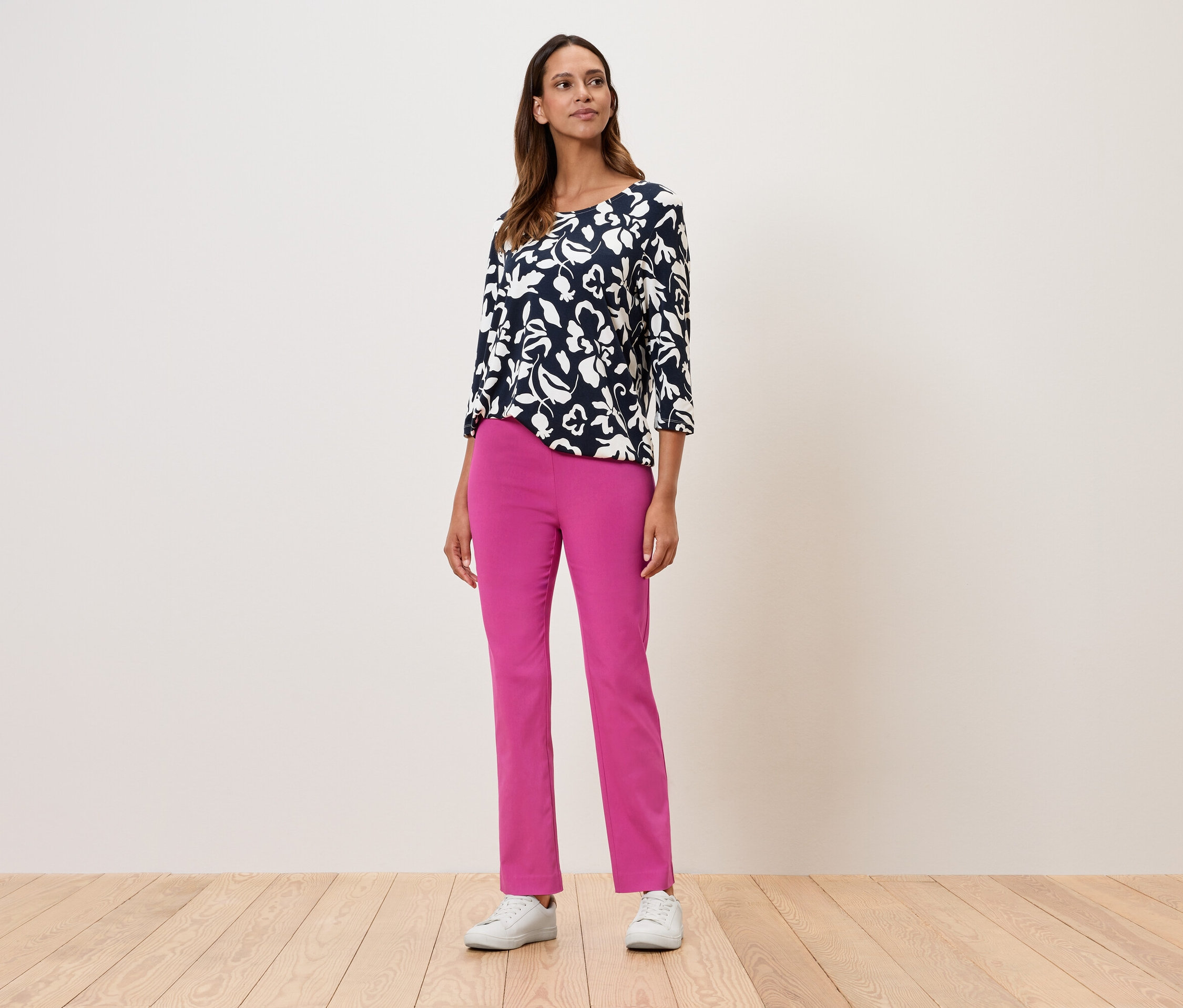 Eine Frau steht und trägt ein geblümtes Oberteil mit Dreiviertelärmeln, eine pinke Hose und weiße Turnschuhe.