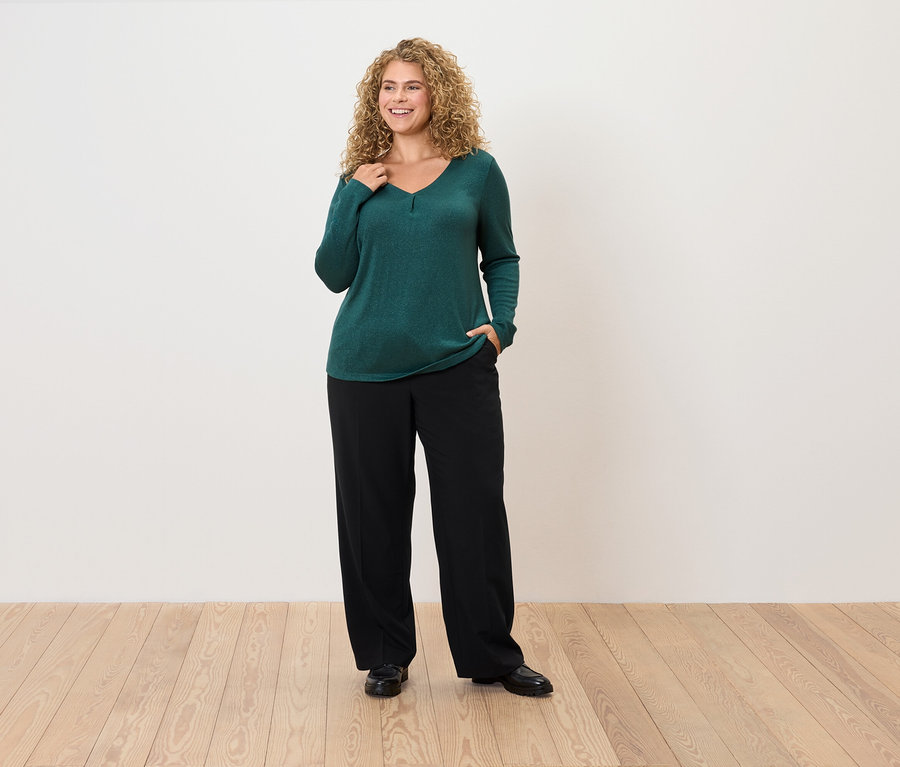 Frau mit lockigem Haar posiert in smaragdgrünem Pullover und schwarzer Hose.
