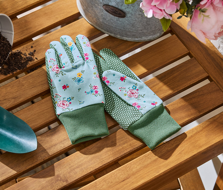 Auf einem Holztisch liegen Gartenhandschuhe mit Blumenmuster, eine kleine Schaufel und ein Topf mit Erde.