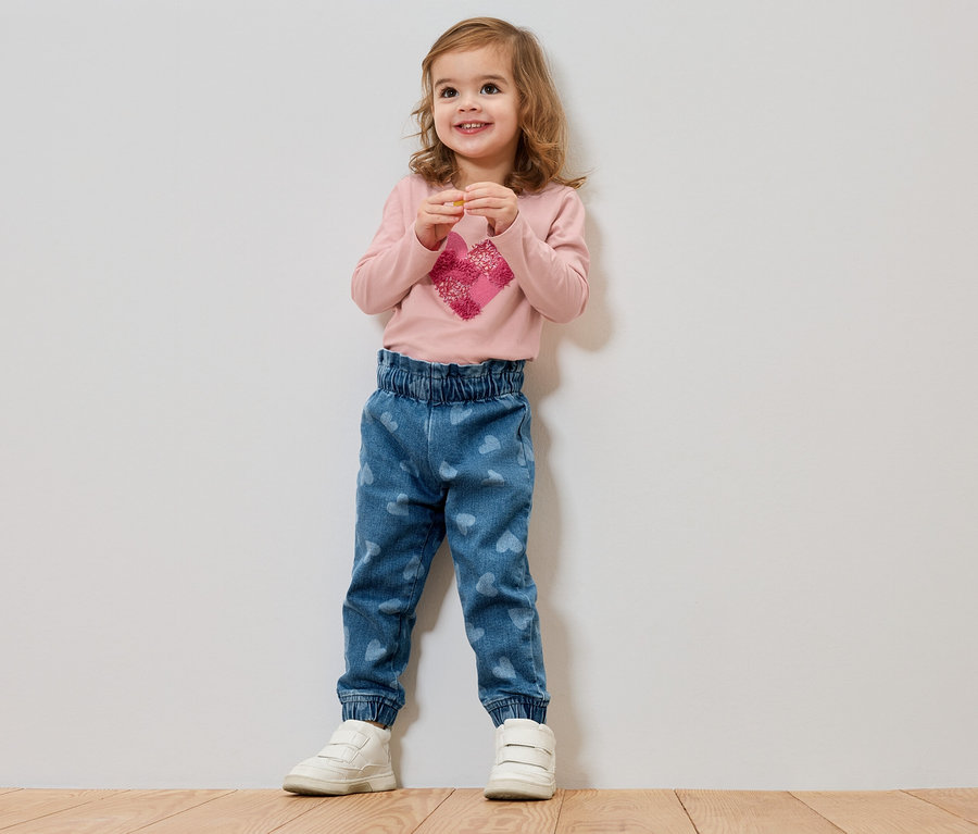 Ein kleines Mädchen mit blonden Haaren steht vor einer Wand und trägt ein rosa Langarmshirt mit einem Herz und blaue Jeans mit Herzen.