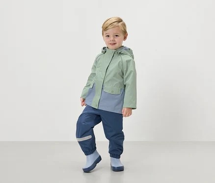 Ein blonder Junge trägt eine mintgrüne Regenjacke mit marineblauen Regenhosen und hellblauen Gummistiefeln.