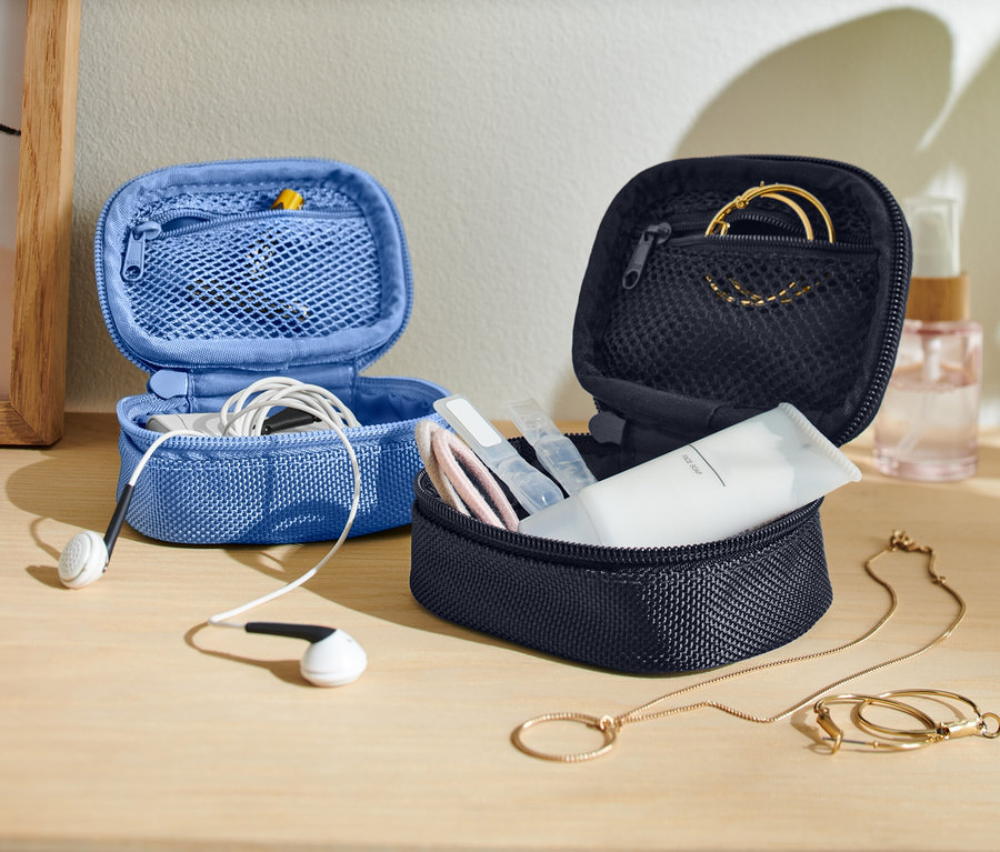 Auf einem Tisch stehen zwei geöffnete, kleine Aufbewahrungstaschen, eine blaue mit Kopfhörern und eine dunkle mit Hautpflegeprodukten und Schmuck.