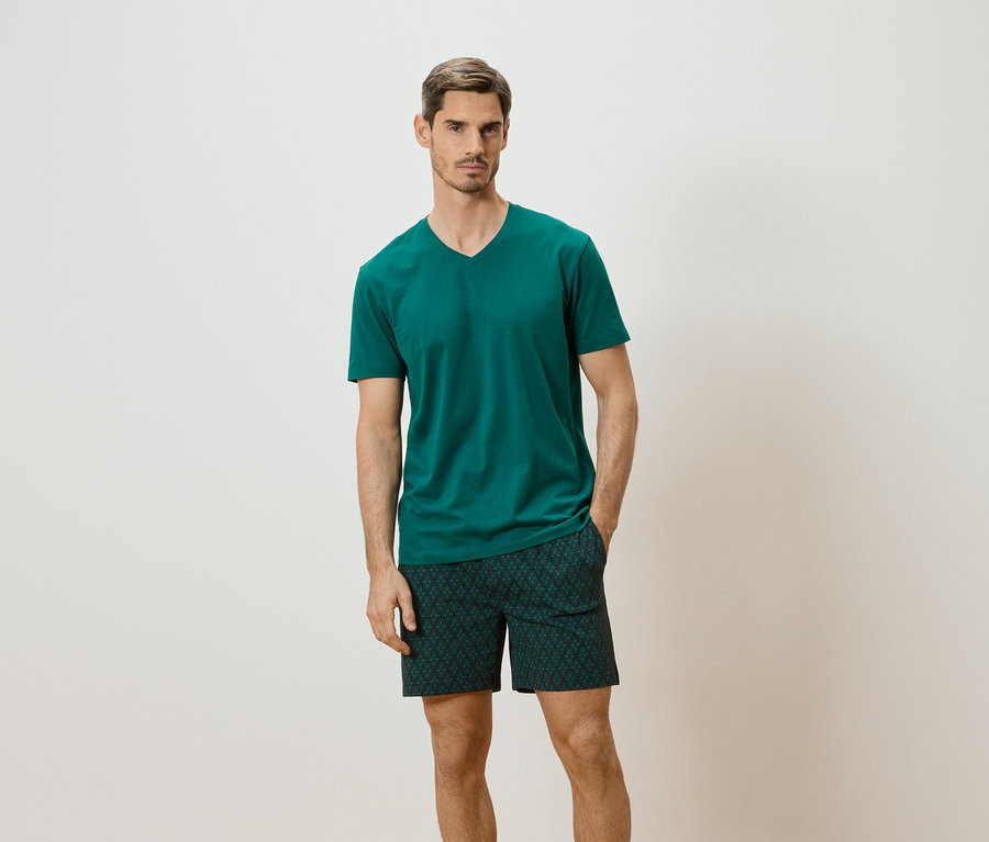 Mann mit smaragdgrünem T-Shirt und gemusterter Shorts vor neutralem Hintergrund.