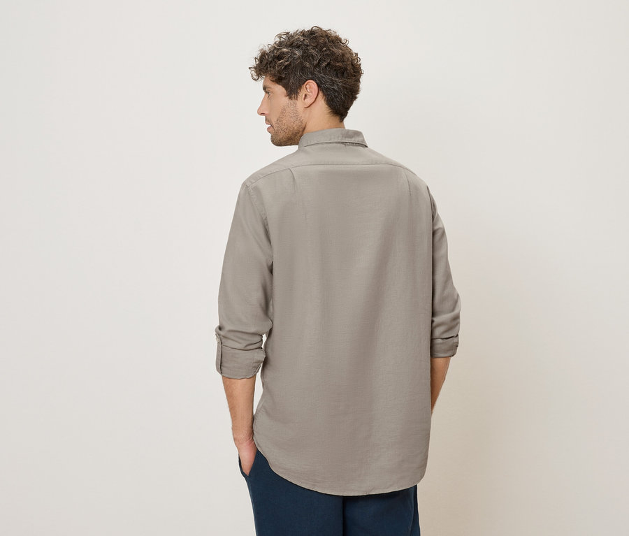 Ein Mann mit lockigem Haar steht mit dem Rücken zur Kamera und trägt ein graues Hemd und eine blaue Hose.