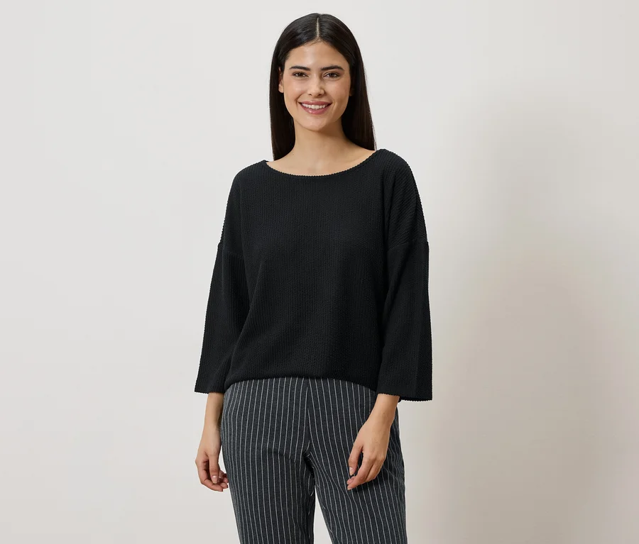 Frau mit dunklen Haaren trägt einen schwarzen Pullover und eine gestreifte Hose vor einem neutralen Hintergrund.
