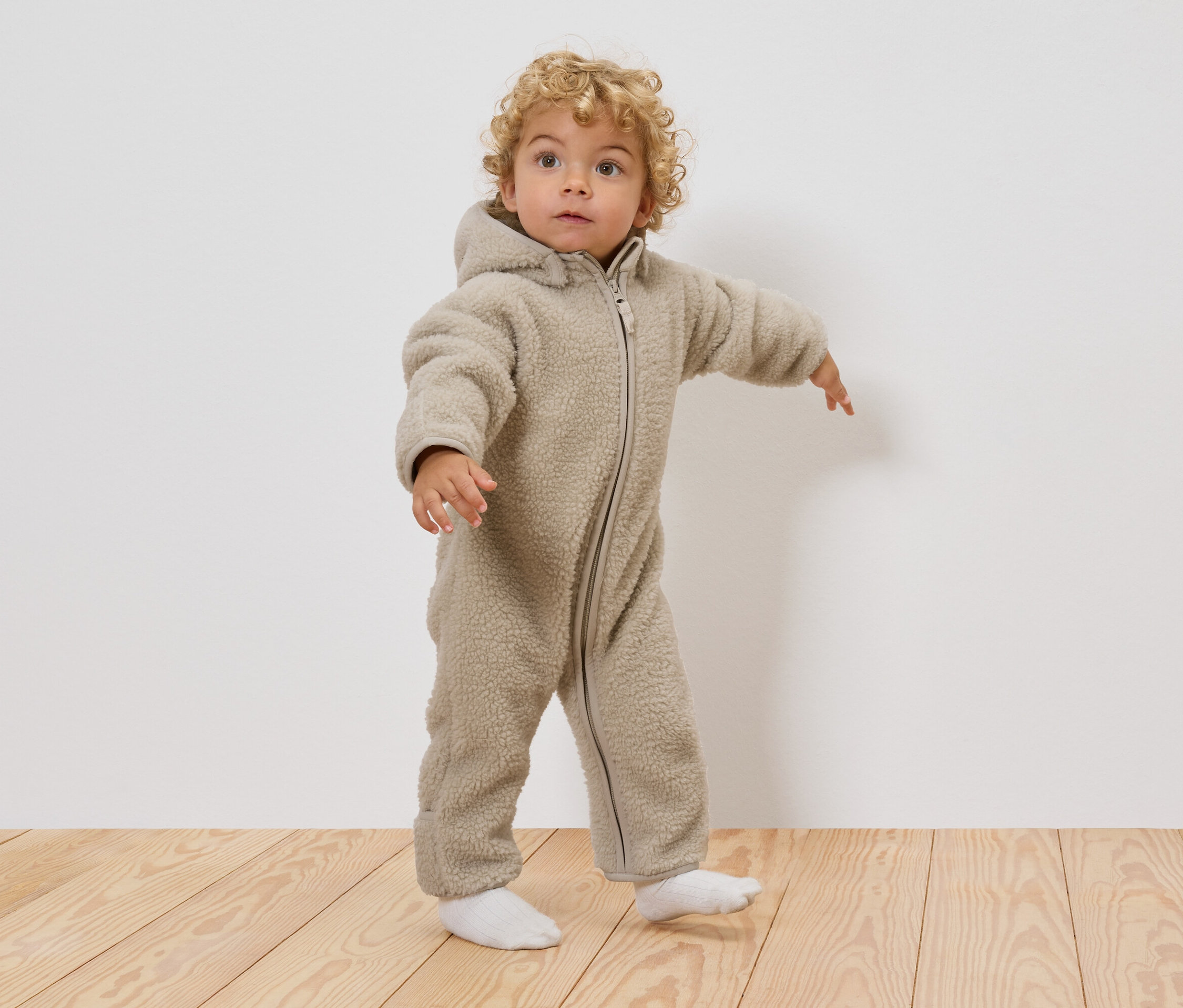 Ein Kleinkind mit lockigen blonden Haaren trägt einen beigen Teddy-Overall mit Kapuze und weißen Socken, während es auf einem Holzboden steht.