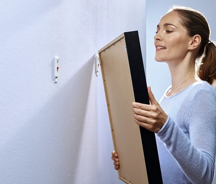 Frau hält ein gerahmtes Bild vor eine Wand mit zwei Klebehaken.