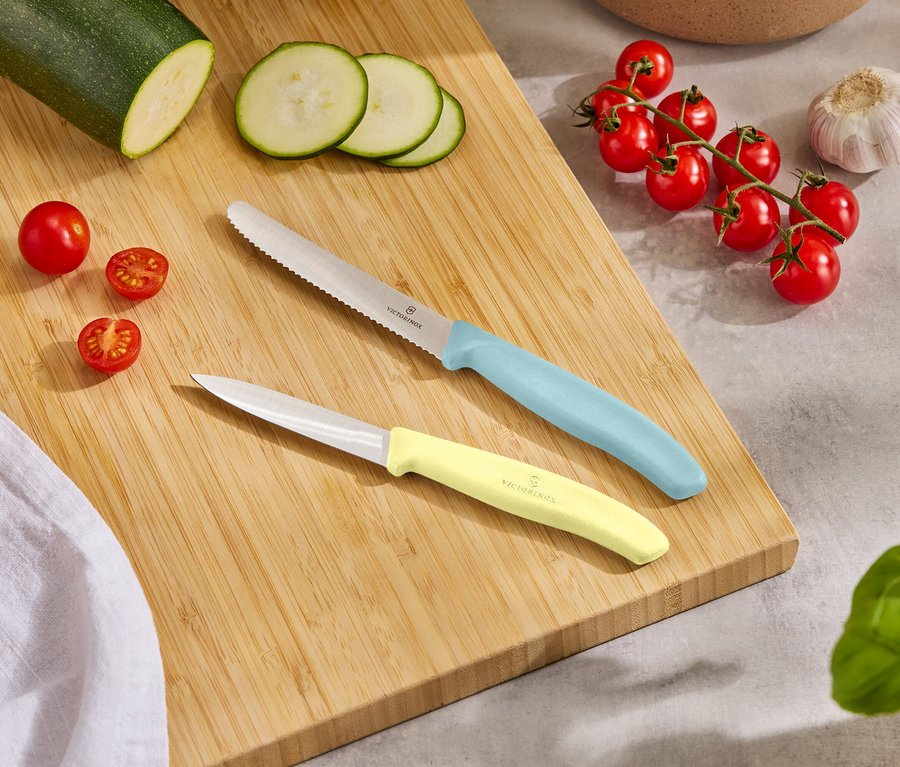 Auf einem Holzbrett liegen ein ganzer Zucchini, Zucchinischeiben, Kirschtomaten, eine Knoblauchzehe und zwei Messer mit hellblauen und gelben Griffen.