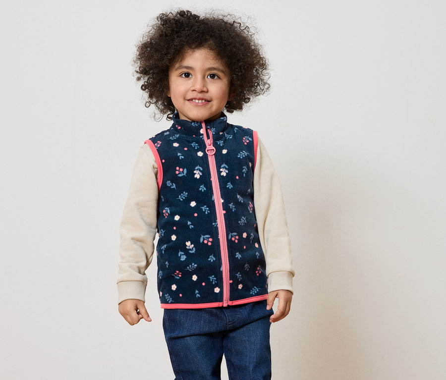 Ein lächelndes Kind mit lockigem Haar trägt eine marineblaue Weste mit Blumenmuster und rosa Reißverschluss über einem beigen Oberteil und Jeans.