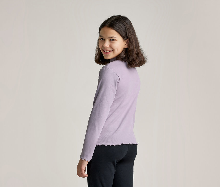 Ein junges Mädchen mit dunklen Haaren, das ein hellviolettes Langarmshirt mit Stehkragen und Rüschen an Ärmeln und Saum trägt, dreht den Kopf, um in die Kamera zu lächeln. Sie trägt auch eine schwarze Hose.