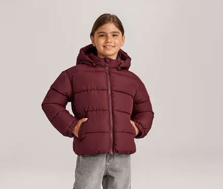 Lächelndes Mädchen in bordeauxroter Steppjacke mit Kapuze und grauer Jeans, die Hände in den Jackentaschen.