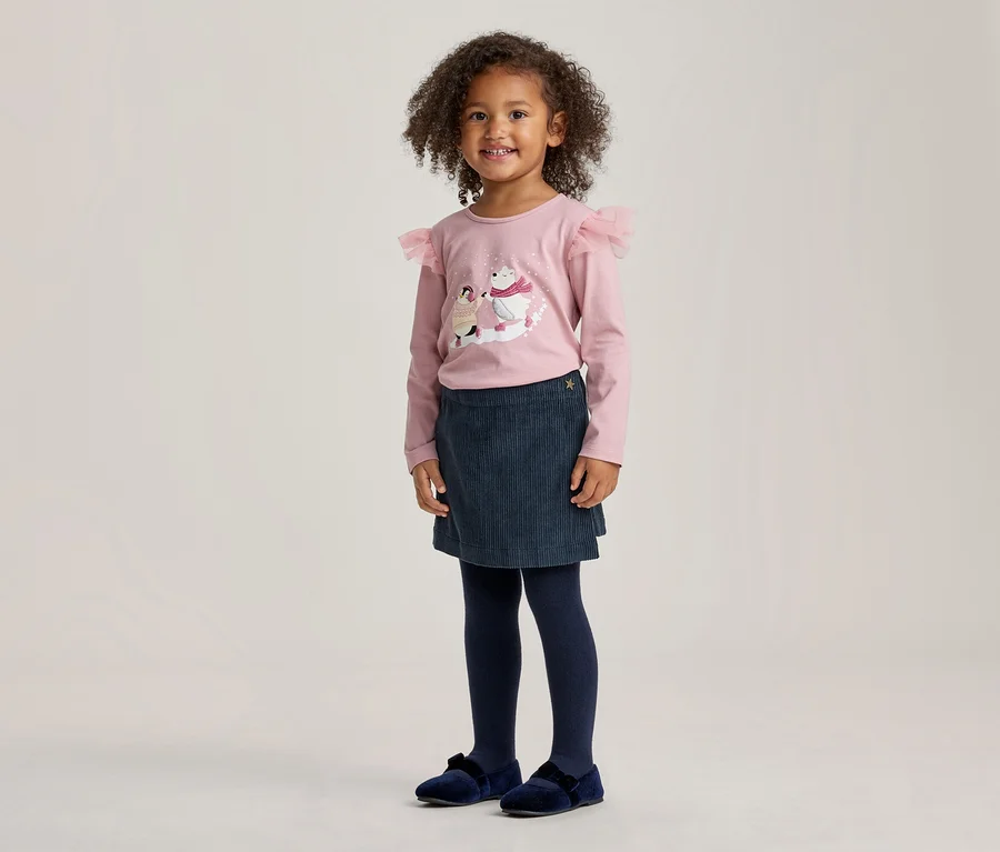 Kleines Mädchen mit lockigem Haar, lächelnd, trägt ein rosa Langarmshirt mit Eisbärenmotiv und Rüschen an den Schultern, einen dunkelblauen Rock, dunkelblaue Strumpfhosen und dunkelblaue Samtschuhe.