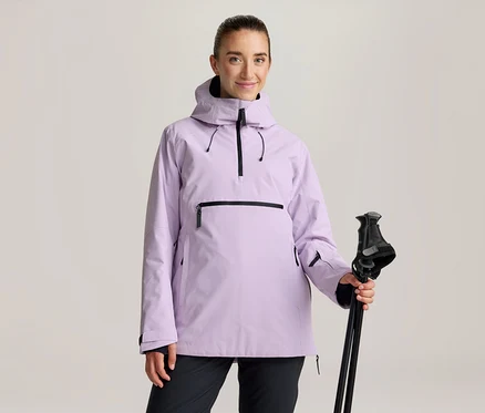 Frau in einer hellvioletten Anorak-Jacke mit schwarzen Reißverschlüssen und schwarzer Hose, die schwarze Skistöcke hält.