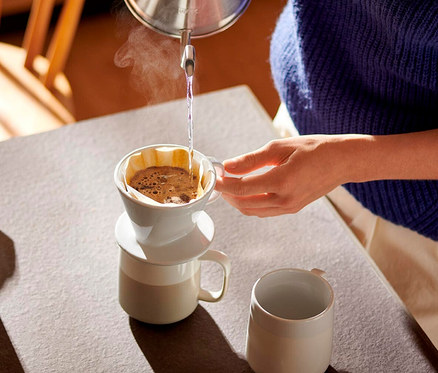 Eine Person gießt heißes Wasser aus einem Wasserkocher in einen weißen Kaffee Handfilter, Größe 101, der auf einer Tasse steht. Dampf steigt aus dem Filter auf. Eine weitere Tasse steht auf dem Tisch.