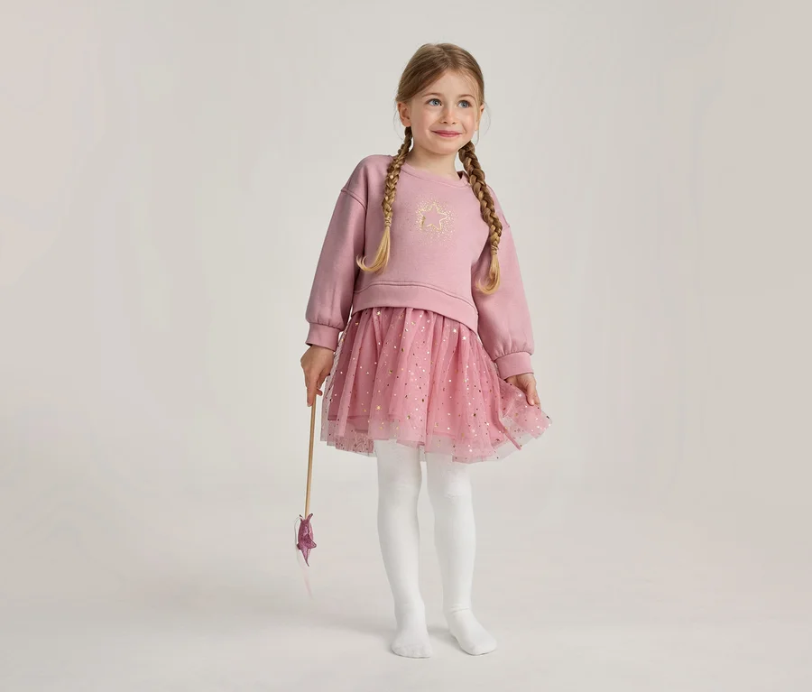 Ein junges Mädchen mit blonden Zöpfen trägt ein rosa Langarmshirt mit goldenem Stern, einen rosa glitzernden Tüllrock und weiße Strumpfhosen, hält einen Sternenzauberstab und lächelt.