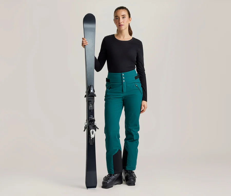 Eine Frau steht mit einem schwarzen Ski. Sie trägt ein schwarzes Langarmshirt, eine türkise Skihose und schwarze Skischuhe.