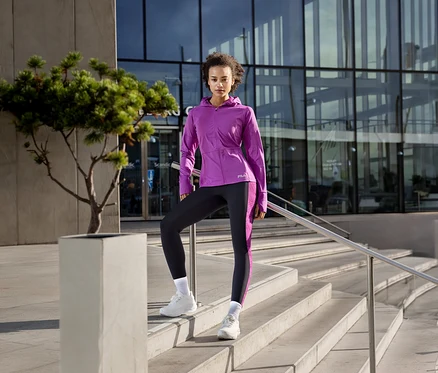 Frau in violetter FILA Damen-Funktionsjacke und schwarzen Leggings steht auf einer Treppe, ein Fuß auf einem Podest.