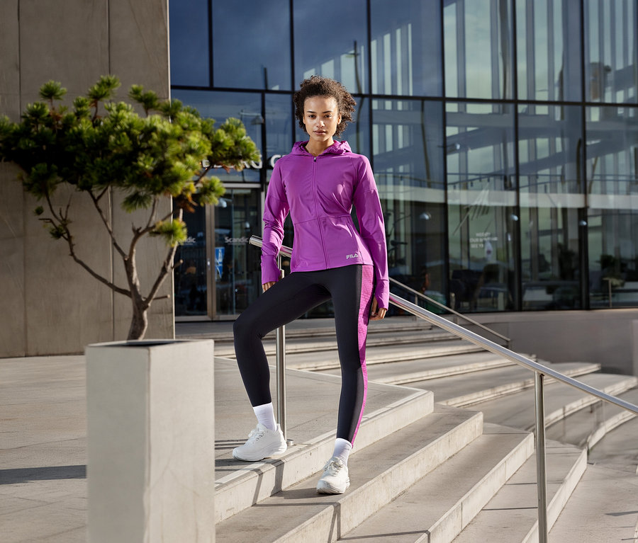 Frau in violetter FILA Damen-Funktionsjacke und schwarzen Leggings steht auf einer Treppe, ein Fuß auf einem Podest.