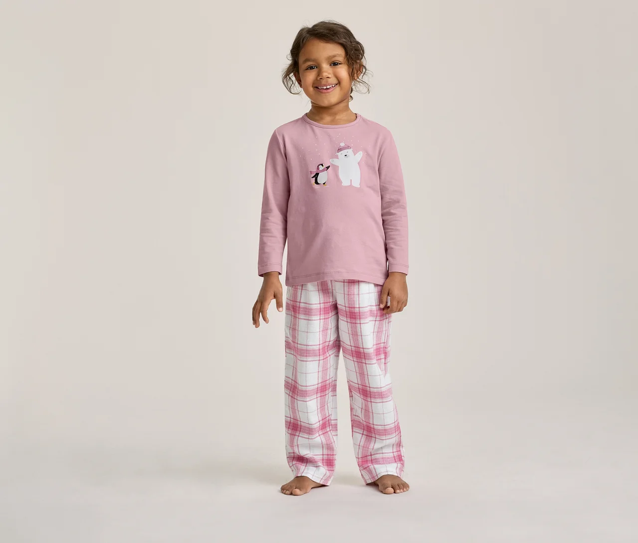 Lächelndes Kind in einem rosa Pyjama-Set mit einem Eisbär- und Pinguin-Aufdruck auf dem Oberteil und rosa-weiß karierten Hosen.