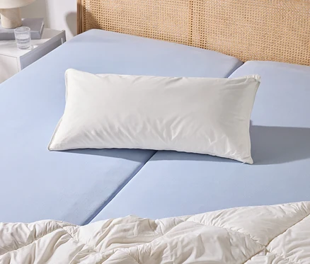 Ein Bett mit hellblauem Spannbettlaken, einem weißen Kissen und einer weißen Bettdecke. Daneben steht ein weißer Nachttisch mit Lampe und Wasserglas.