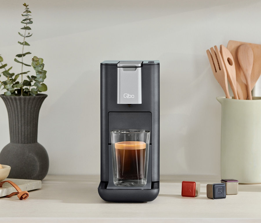 Eine Qbo ESSENTIAL Dark Stone Kaffeemaschine mit einer Tasse Kaffee. Daneben stehen Kapseln und eine Schale mit Himbeeren.
