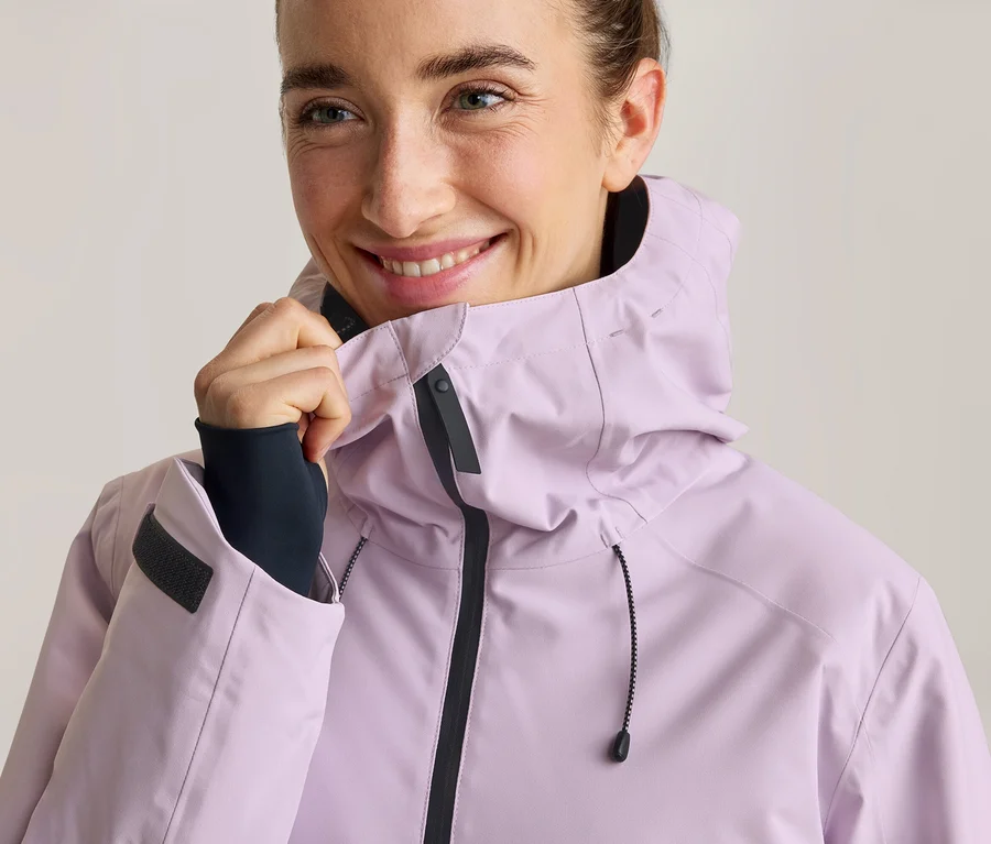 Lächelnde Frau in einer hellvioletten Kapuzenjacke, die den Kragen hält. Sie trägt ein dunkles Langarmshirt unter der Jacke.