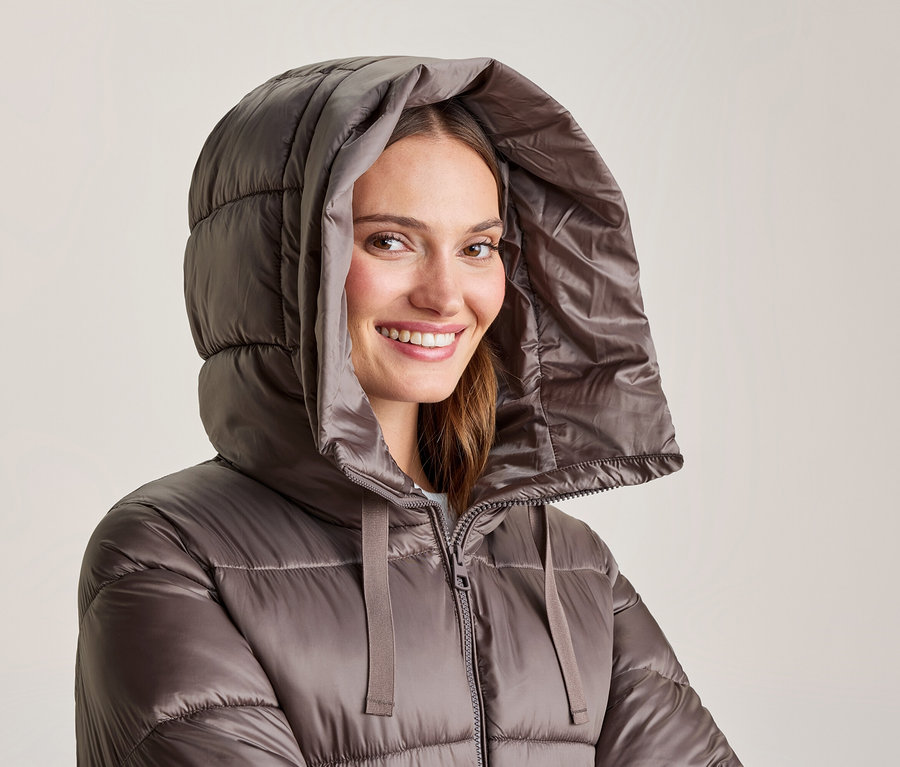 Lächelnde Frau in glänzender brauner Steppjacke mit hochgezogener Kapuze.