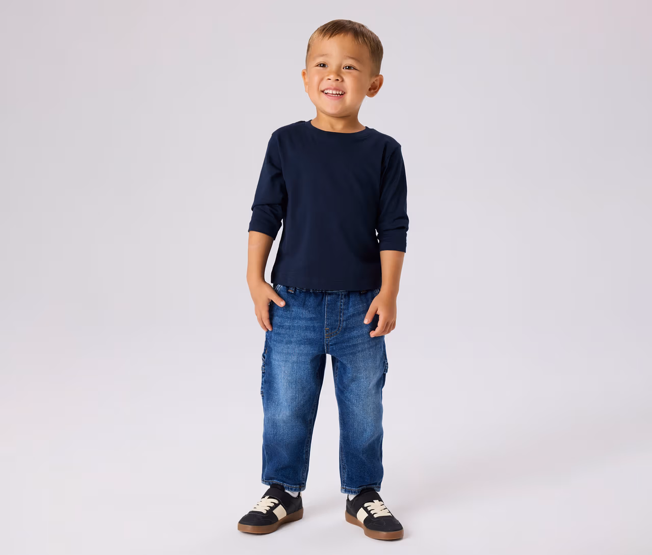 Junger Junge lächelt, trägt ein dunkelblaues Langarmshirt, blaue Jeans und schwarze Sneaker mit weißen Streifen.