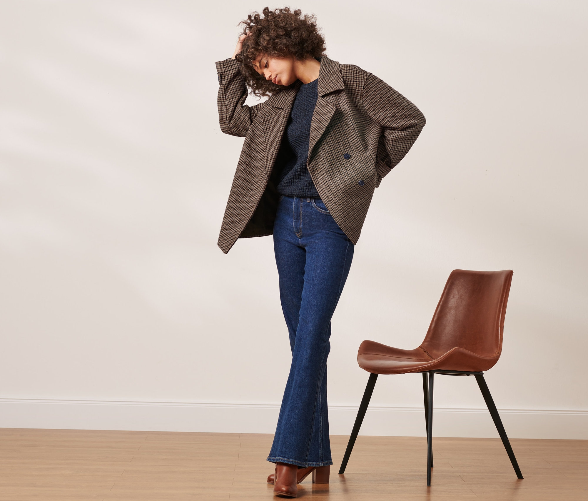 Frau posiert in karierter Jacke mit Wolle, Pullover, Jeans und Schuhen neben einem braunen Stuhl.