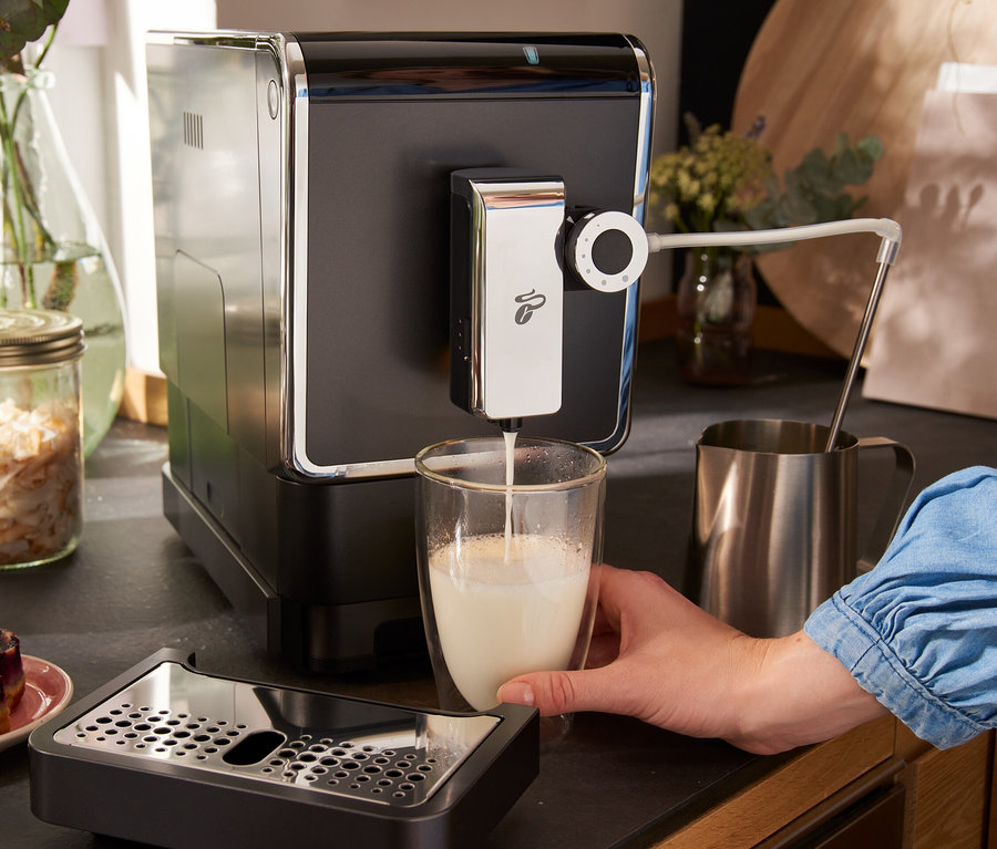 Frau füllt Milch aus einem »Esperto Pro« Tchibo Kaffeevollautomat, Anthrazit, in ein Glas. Auf dem Tisch stehen auch »Esperto Pro« Tchibo Kaffeevollautomaten in Dark Red und Metallic Mint.