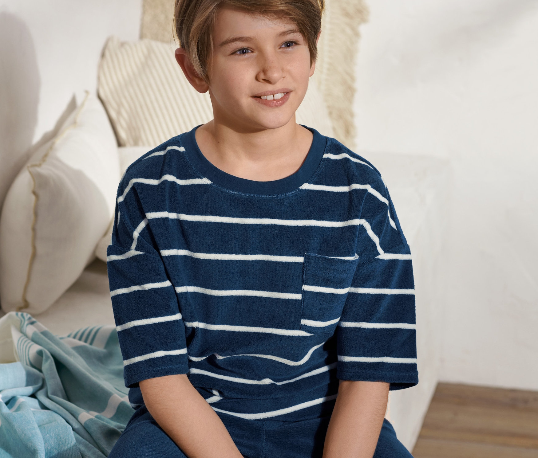 Ein Junge sitzt auf einem Bett und trägt ein blau-weiß gestreiftes Frottier-Shirt.