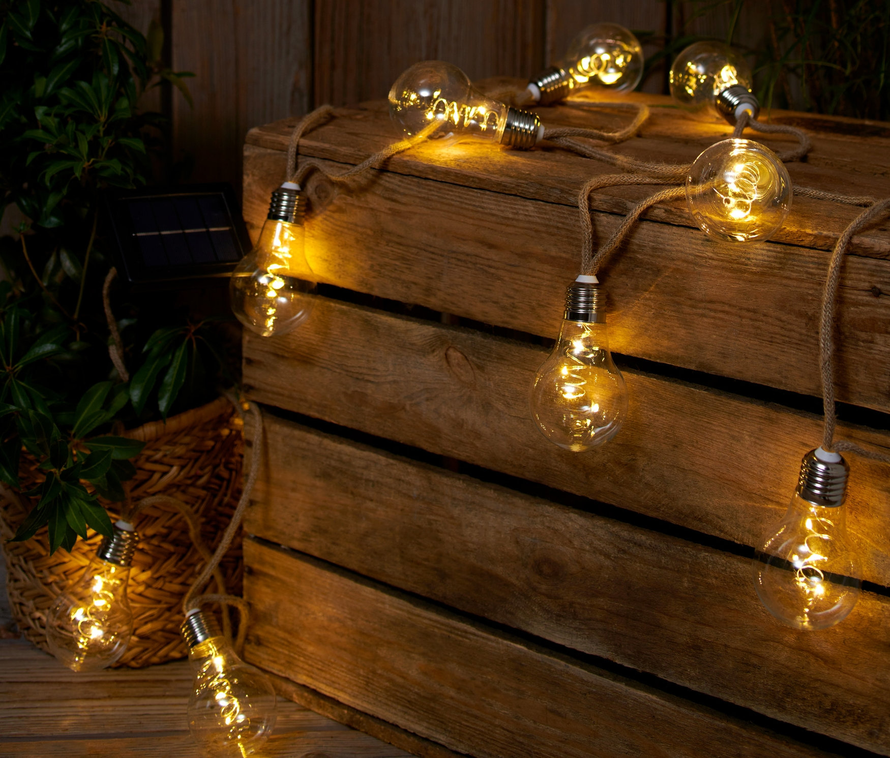 Eine Solar-Lichterkette mit Juteseil liegt auf einer Holzkiste neben einer Pflanze.
