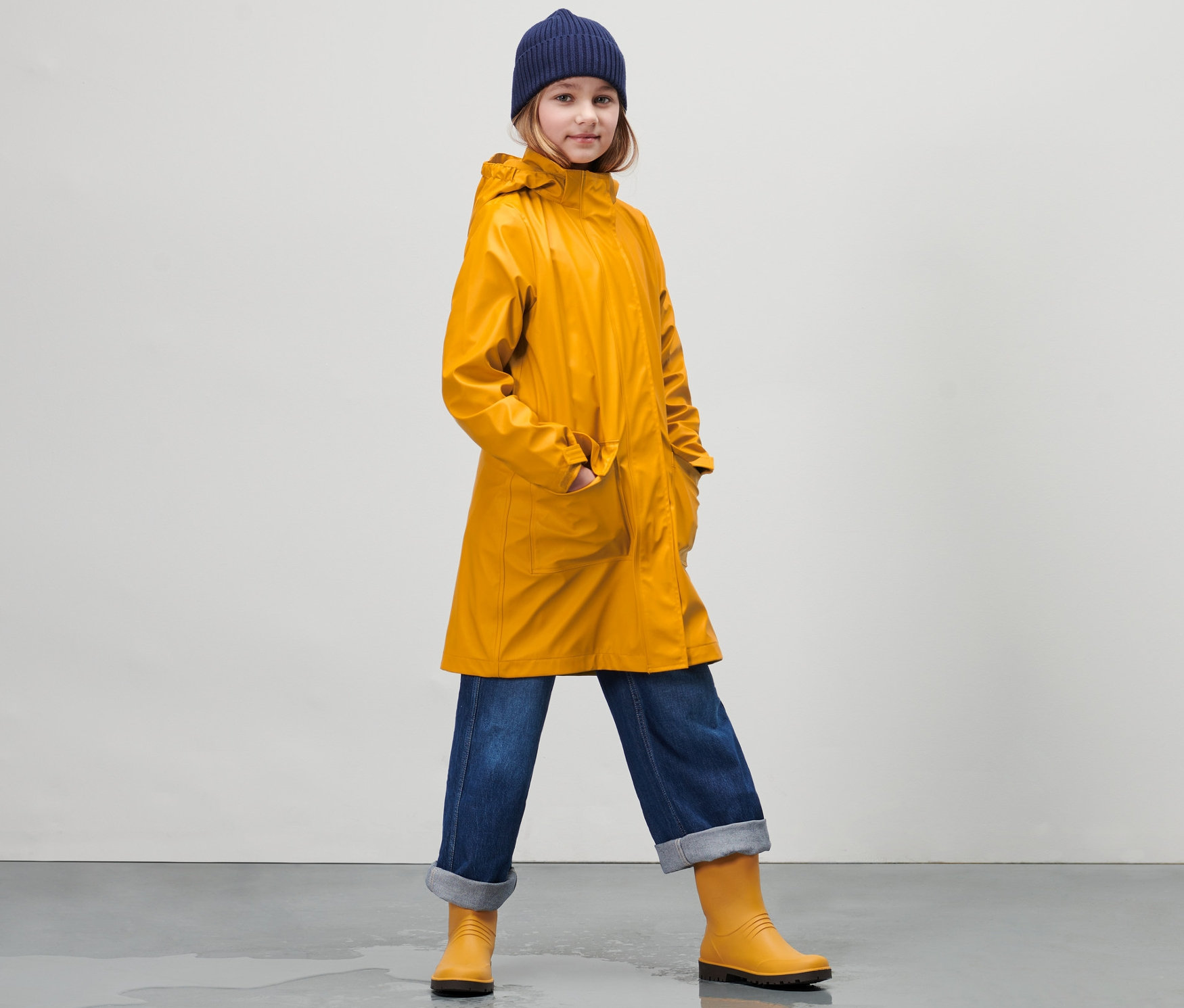 Mädchen in Kinder-Regenparka »Friesennerz«, Jeans und Gummistiefeln steht auf nassem Boden.