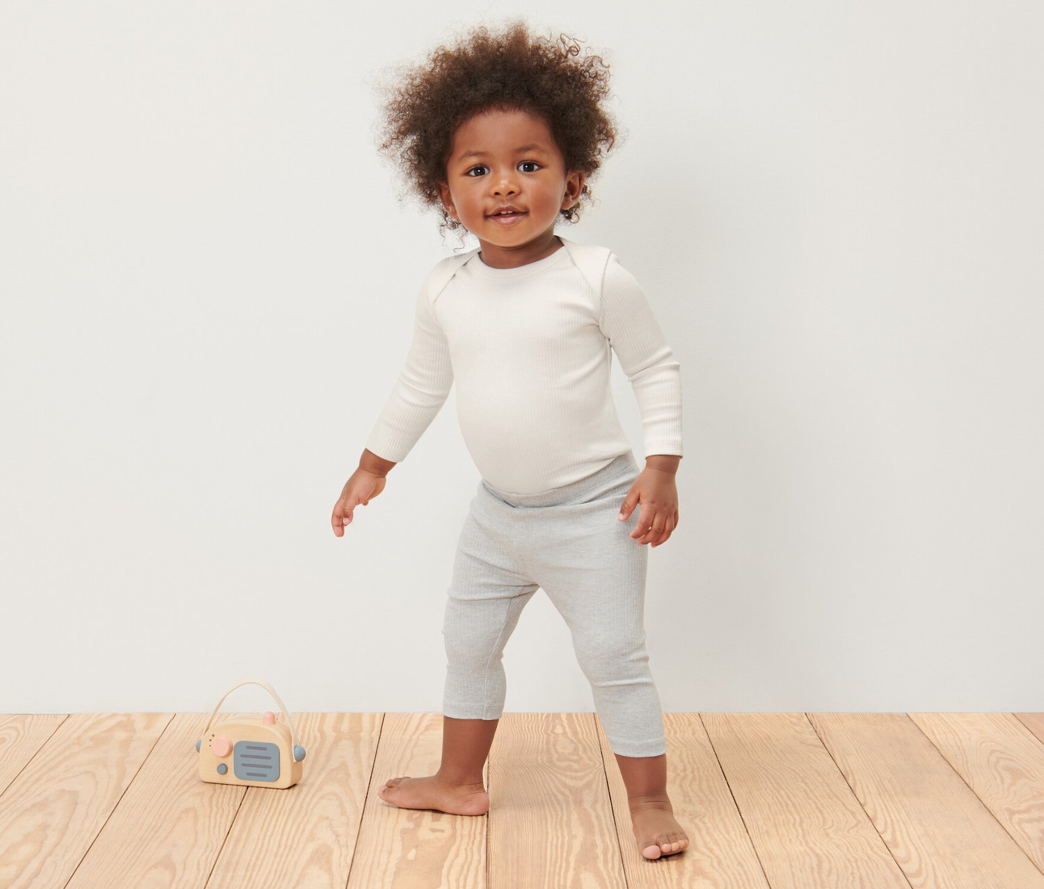 Ein Kind steht auf einem Holzboden und trägt 3 Baby-Ripp-Langarmbodys und graue Leggings.