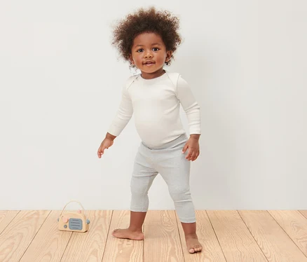 Ein Kind steht auf einem Holzboden und trägt 3 Baby-Ripp-Langarmbodys und graue Leggings.