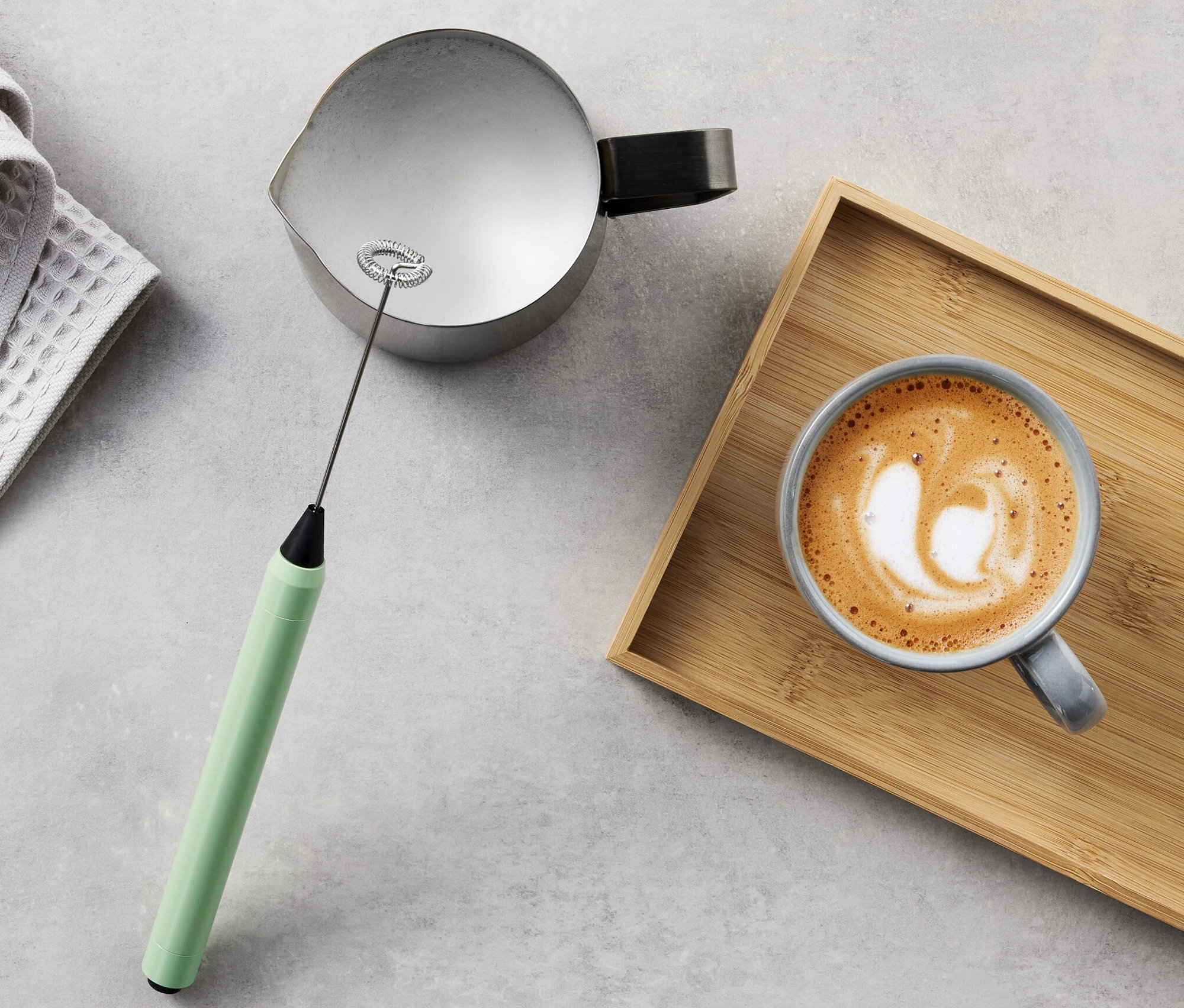 Graues Tuch, Kanne, Milchaufschäumer und Milchkaffee auf Holztablett.