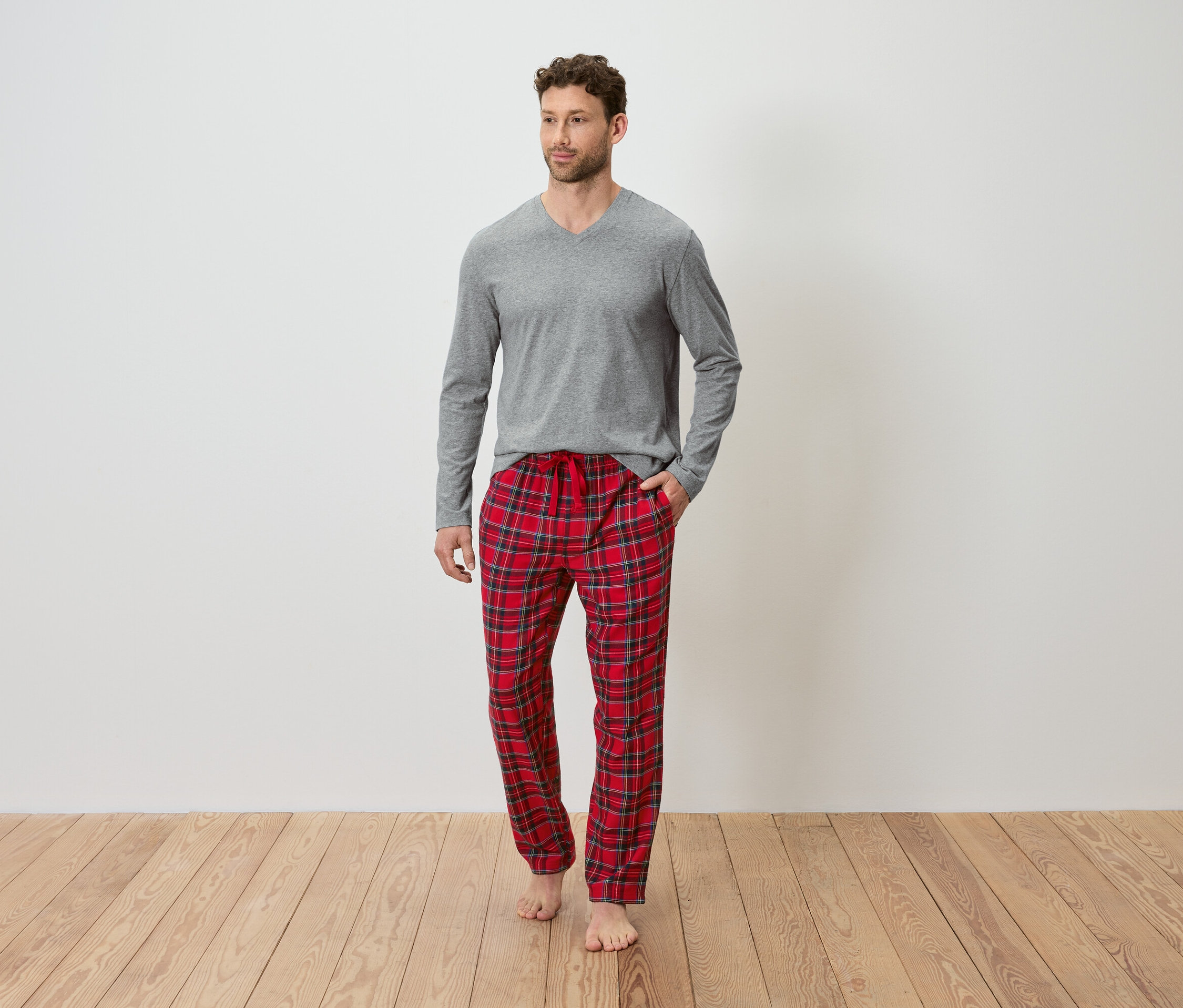 Mann mit grauem Langarmshirt und rot karierter Pyjamahose steht auf Holzboden.