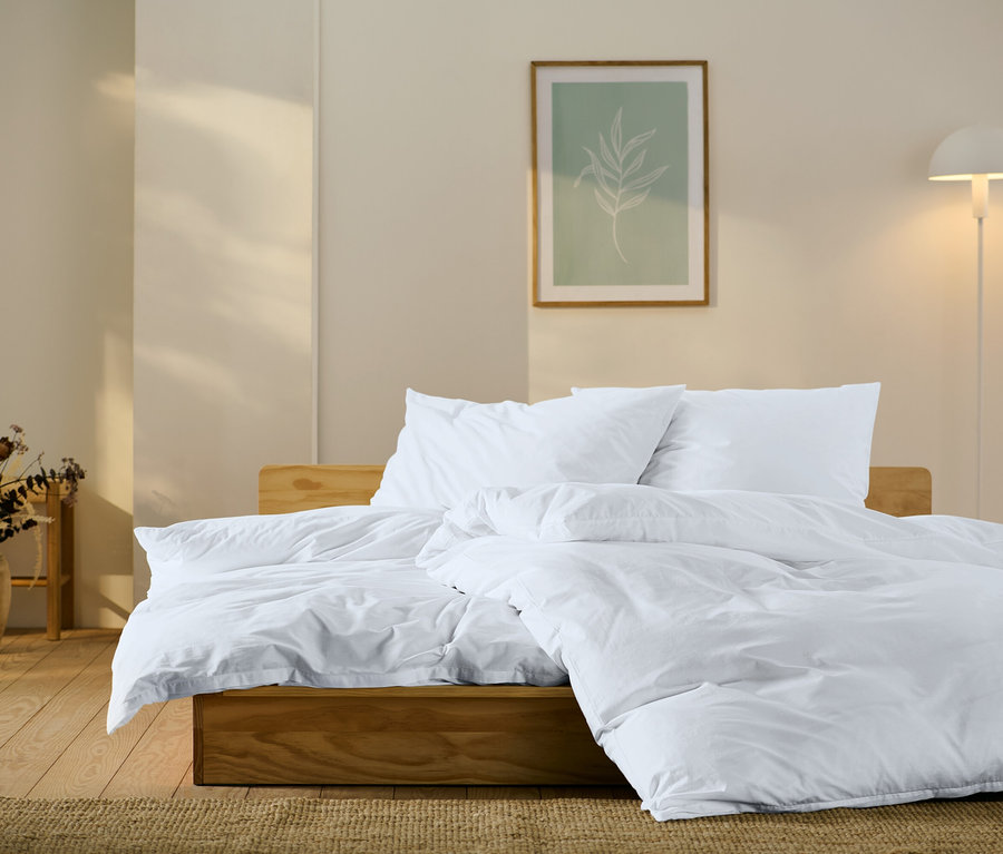 Holzbett mit weißer Bettwäsche, Kissen und Bettdecke. An der Wand ein Bild mit einem Blattmotiv und eine Lampe.