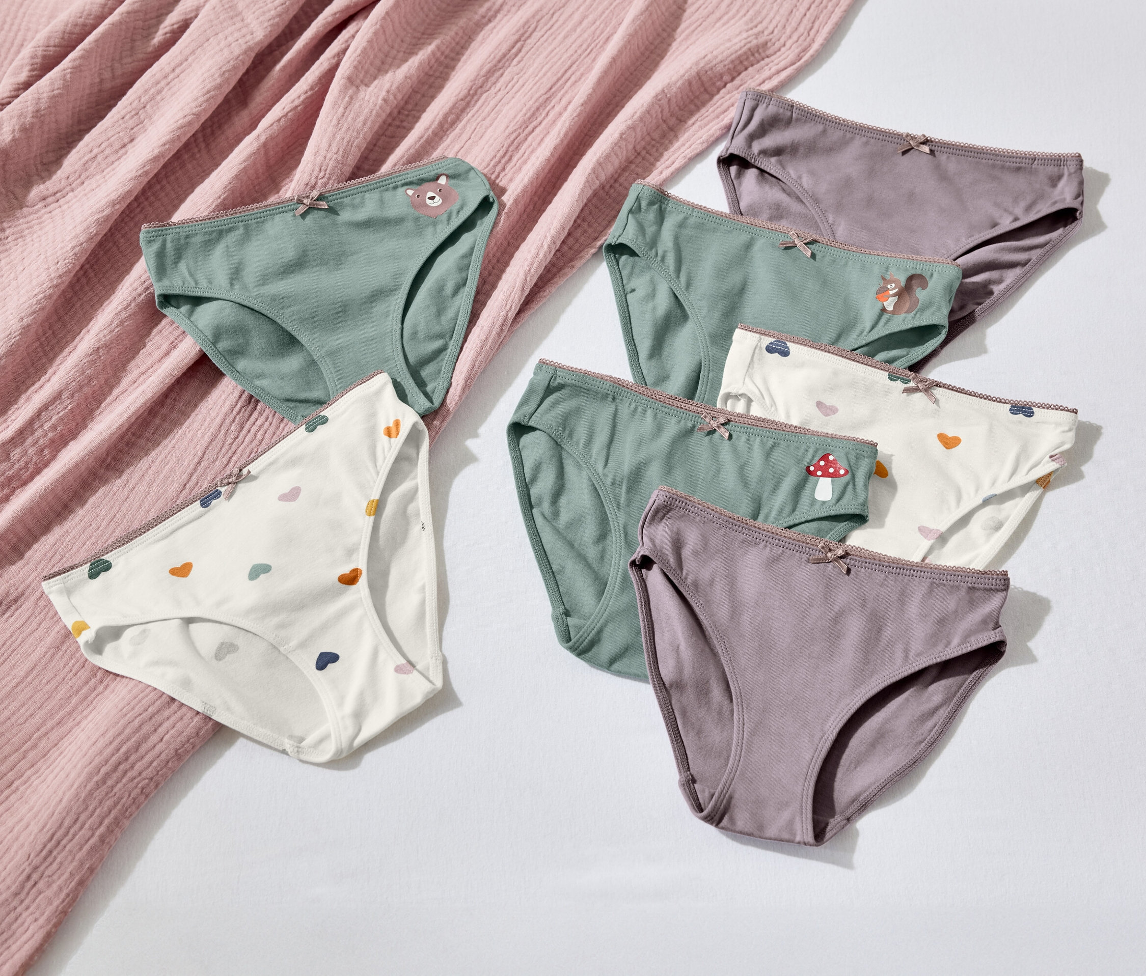 Sieben Paar Damenunterwäsche in verschiedenen Farben und Mustern liegen auf einem weißen Hintergrund neben einem rosa Stoff.