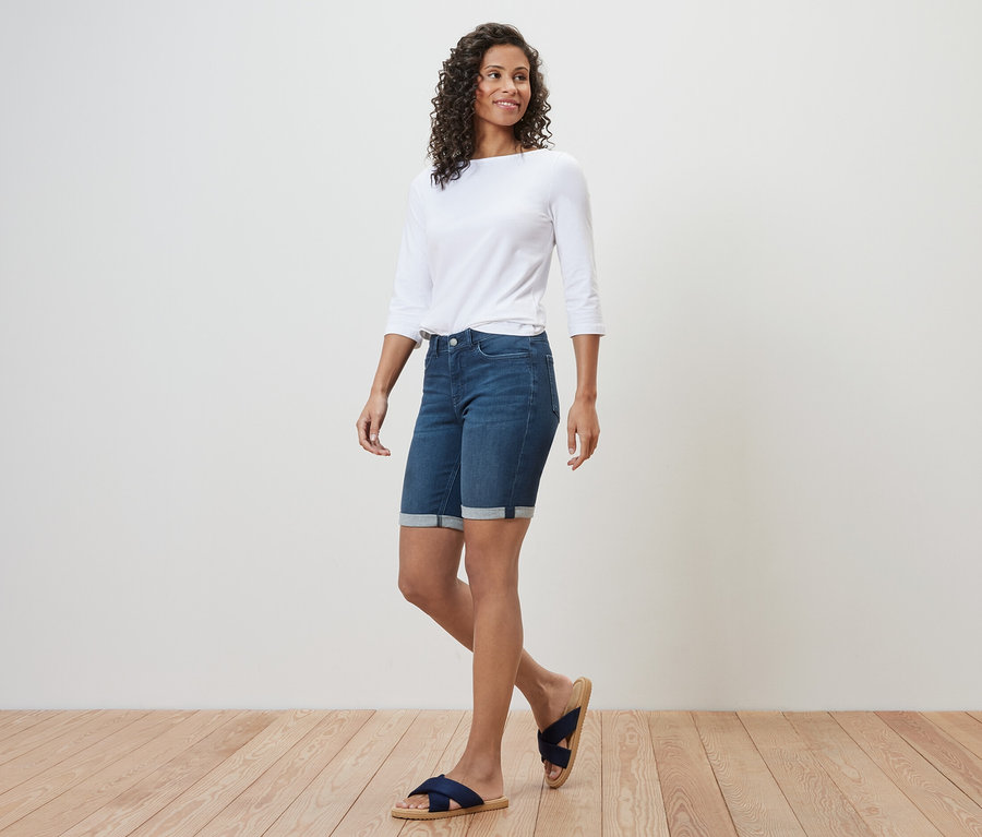 Frau geht, trägt Bermuda-Jeans ‒ Fit »Lea«, dark blue Denim, ein weißes Dreiviertelarm-Top und Sandalen.