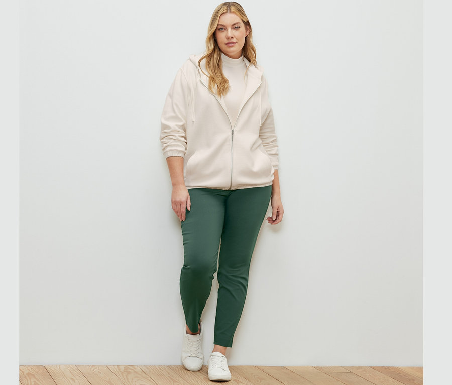 Frau posiert in cremeweißer Sweatjacke, Stretchhose und Blusenshirt mit Stehkragen.