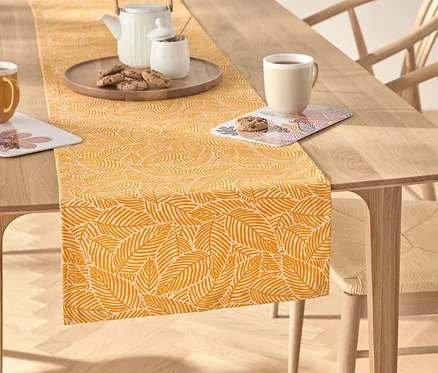 Auf einem Holztisch liegt ein gelber Tischläufer mit Blattmuster, auf dem sich ein Teeservice, eine Tasse und Kekse befinden.