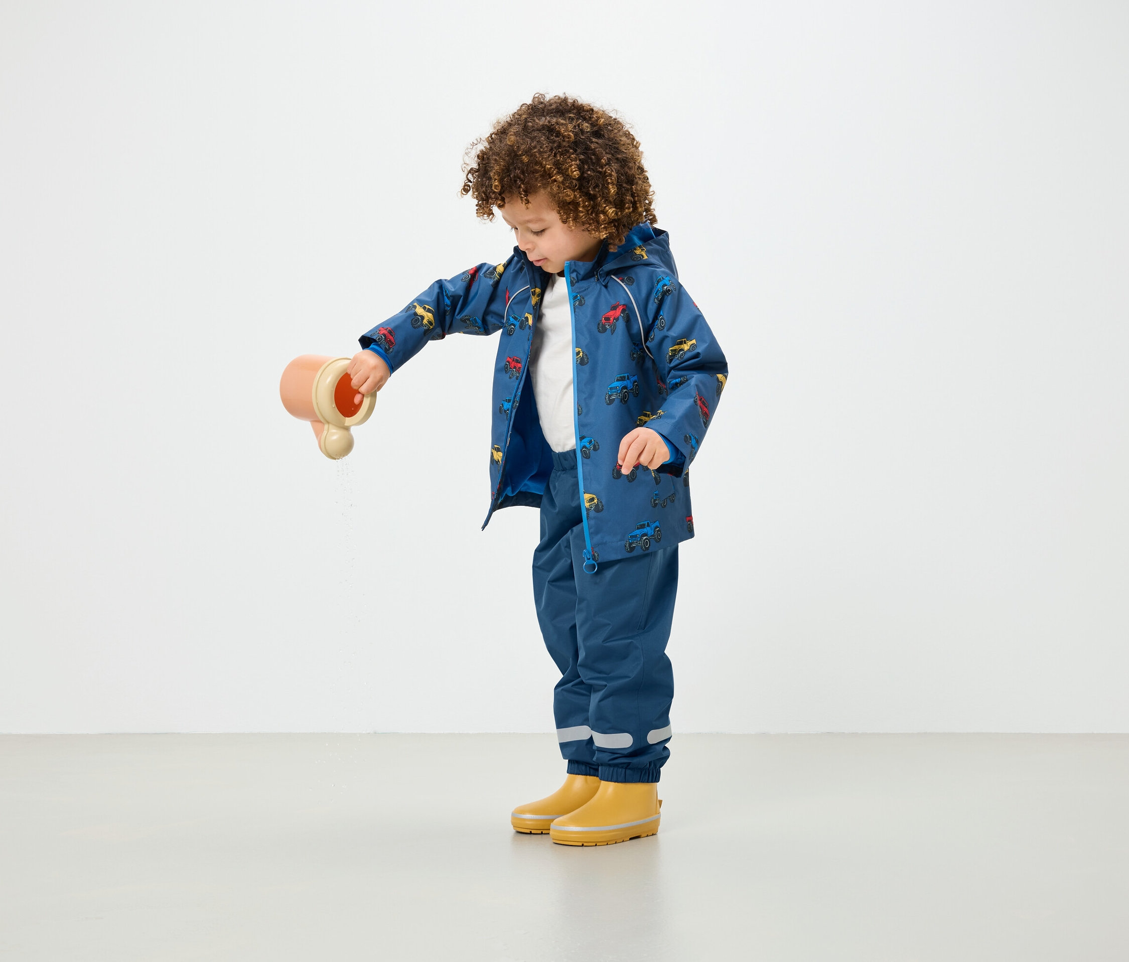 Ein Kind mit lockigem Haar gießt Wasser aus einem Spielzeug, während es einen blauen, bedruckten Regenmantel und blaue Hosen trägt.