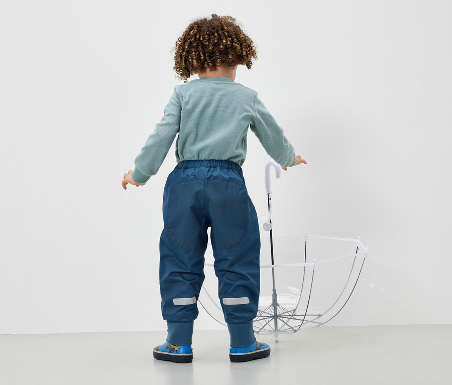 Ein Junge in blauen Hosen und blauen Stiefeln steht mit dem Rücken zur Kamera und hält einen weißen Regenschirm.