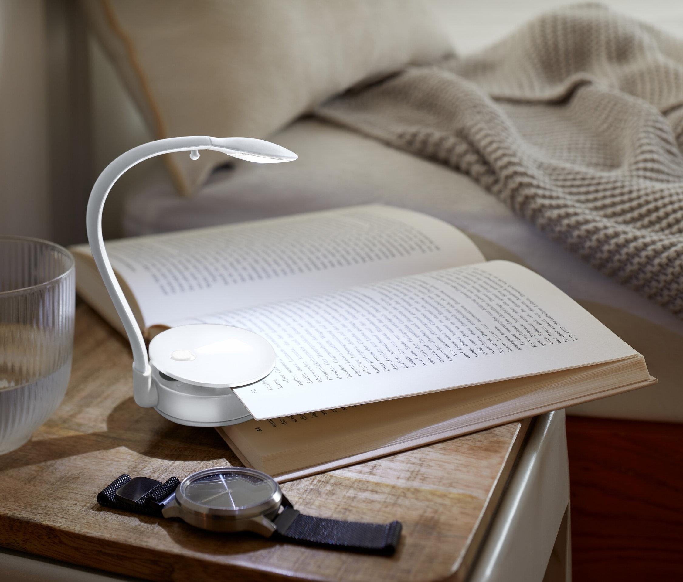 Auf einem Nachttisch liegen ein offenes Buch, das von einer kleinen Lampe beleuchtet wird, daneben ein Glas Wasser und eine Uhr.