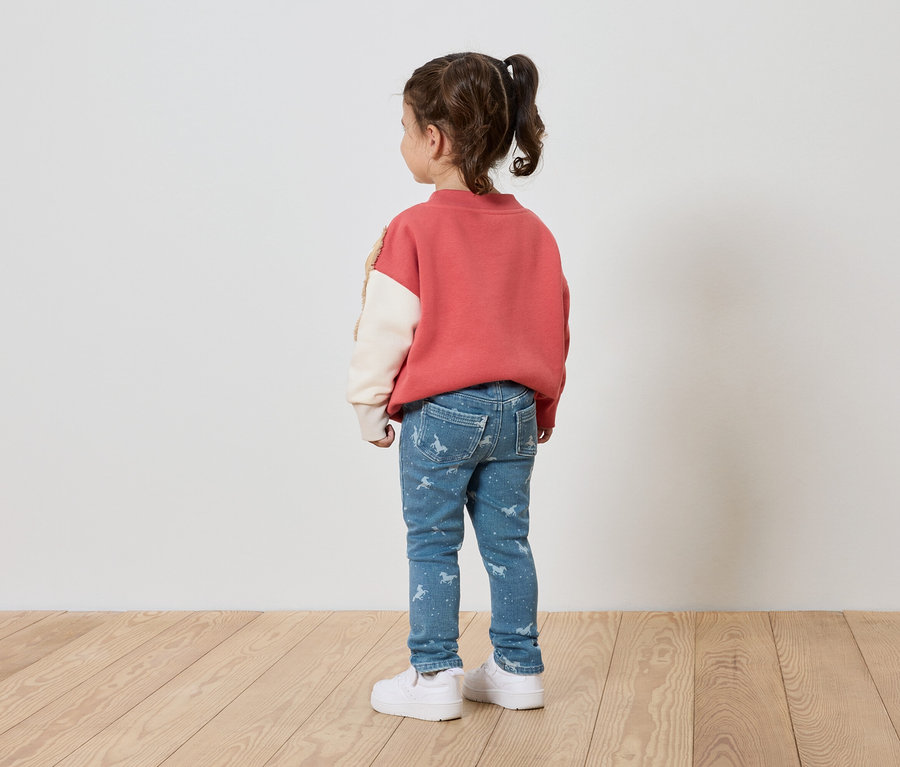 Mädchen mit rosa Pullover, blauen Jeans mit Aufdruck und weißen Schuhen steht mit dem Rücken zur Kamera.