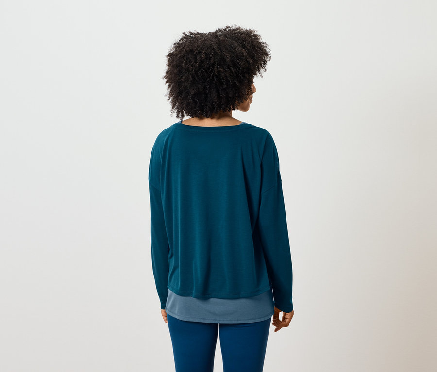 Rückansicht einer Frau mit Afro-Frisur, die eine blaugrüne, langärmlige Bluse und eine blaue Hose trägt.